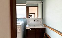 洗面室リノベーション後の写真。ダークブラウンの洗面台と落ち着いたグリーンの壁紙、手すり付きで安心の空間。